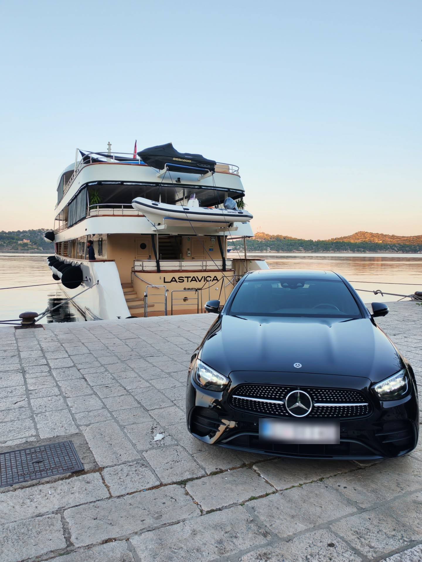A black saloon car parked on a stone quay beside a moored cruise boat at dawn